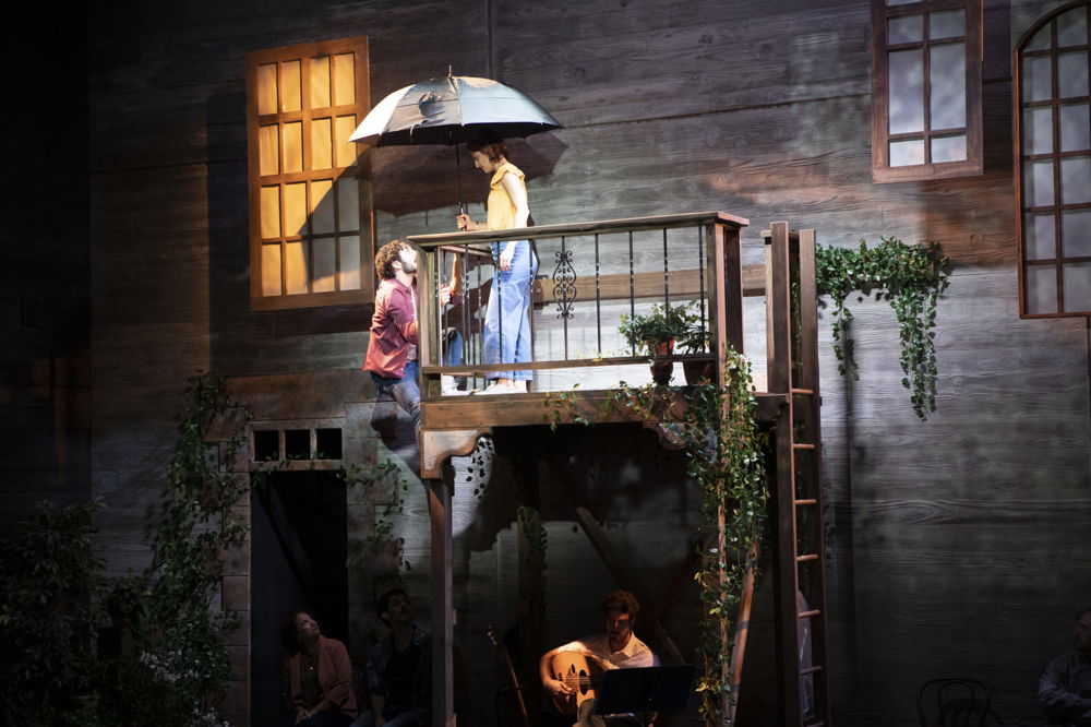 On a stage set, a man climbs a balcony while a woman with an umbrella stands at the top, looking down at him. The walls of the set are decorated with ivy and hanging windows.