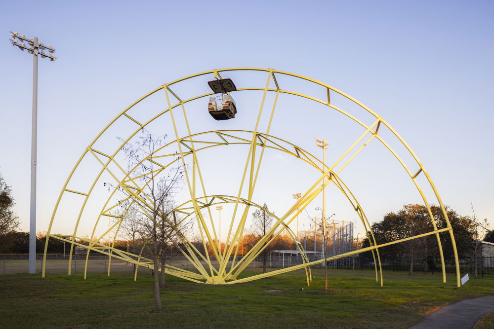 A sculpture resembling the framework of a small, yellow Ferris wheel that has been halved into a semicircle, giving the illusion that half of the ride has been swallowed up by the green lawn below. All of the Ferris wheel’s carriages have been removed except for one at the very top of the ride.