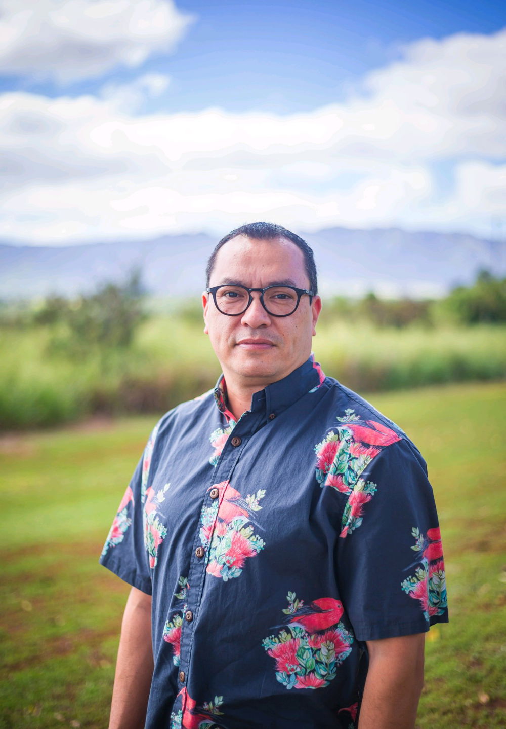 Kawika stands against the Ewa/Waipio plains on Oʻahu. He is wearing a ʻIʻiwi aloha shirt and dark glasses.
