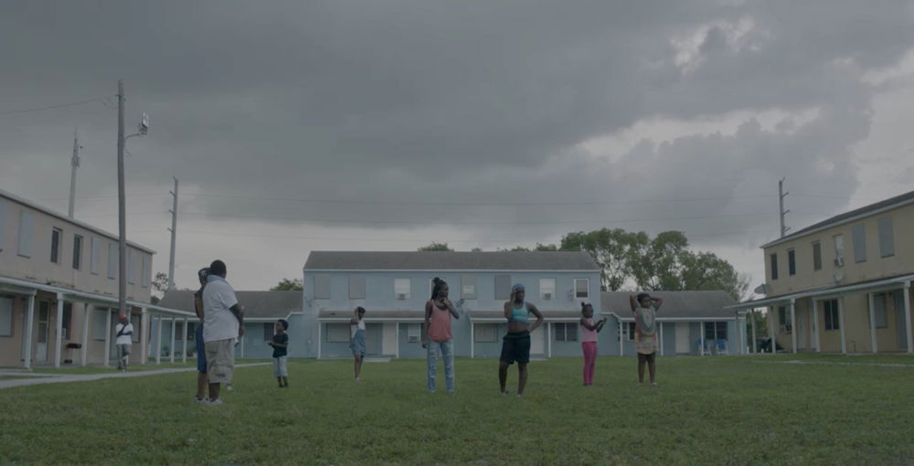 Black people stand together in a vast, open terrace under cloudy skies and flanked by boarded row houses.