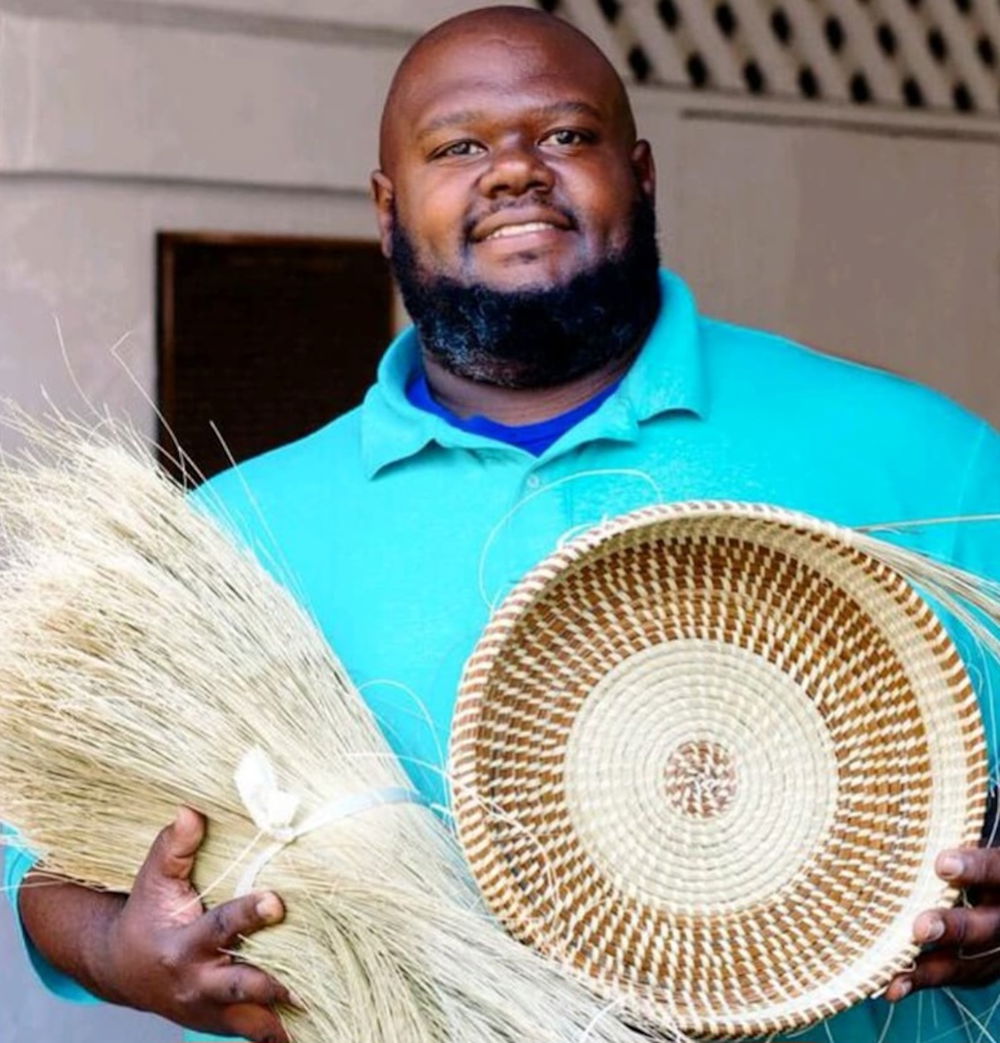 The artist Corey Alston, a man with brown skin, a bald head, and a full beard smiles at us. He has small dimples on both cheeks. He is seen from the waist up holding an unfinished woven basket in his left hand and a bundle of sweetgrass in his right. He wears a robin's-egg-blue collared shirt with an electric-blue undershirt.