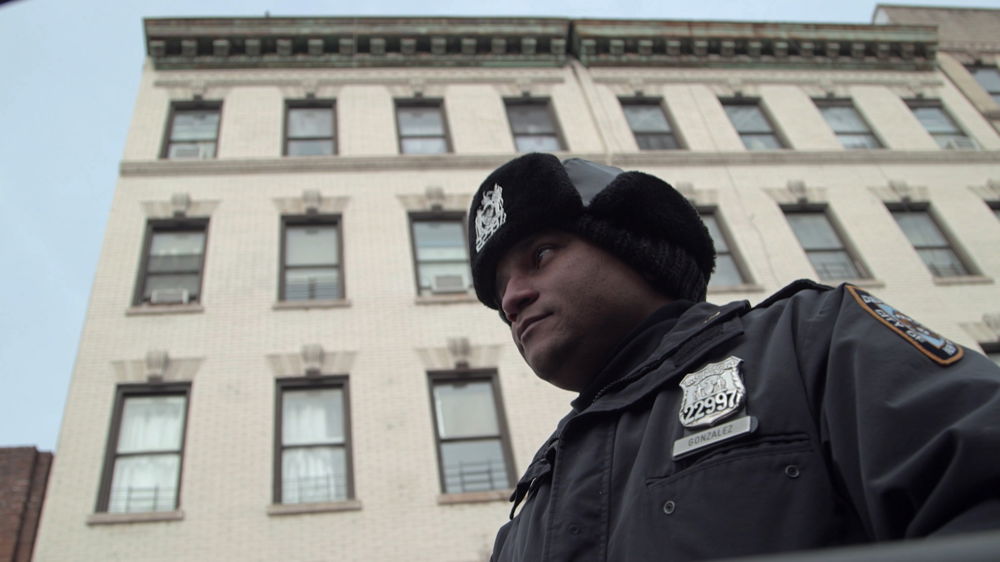 Framed at a low angle in front of an apartment building, an NYPD cop in a winter hat looks warily into the distance.