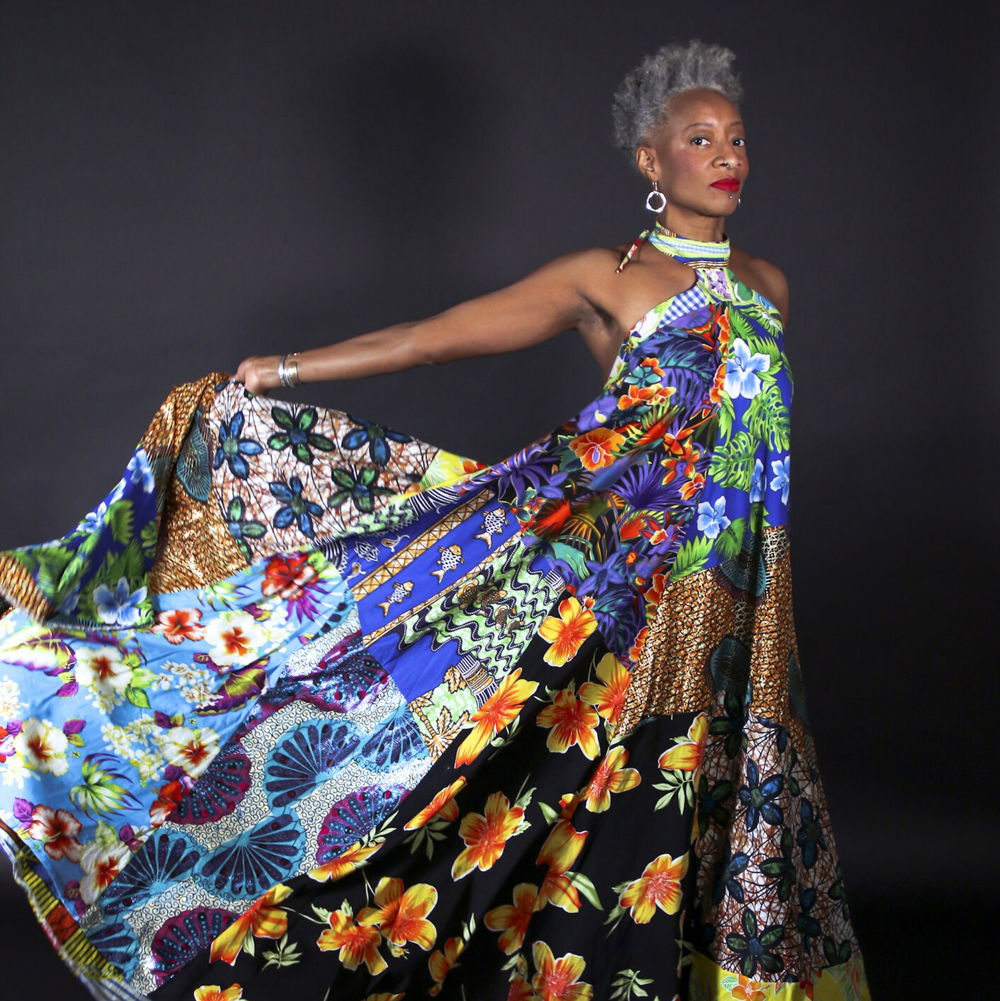 Cynthia stands in profile extending an arm behind her to hold and display the skirt of her long, flower-printed dress designed and crafted by Robert Young.