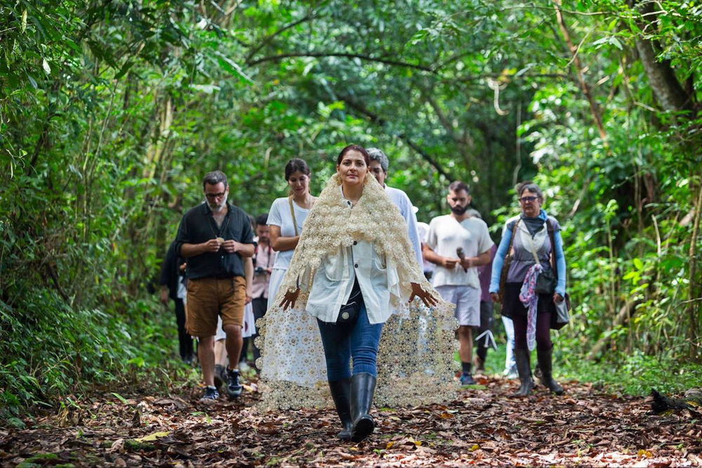 A photograph of a group of people walking toward the camera on a pathway covered with fallen leaves. They are in a lush, green wooded space. A woman wearing a lacy white shawl leads the group.