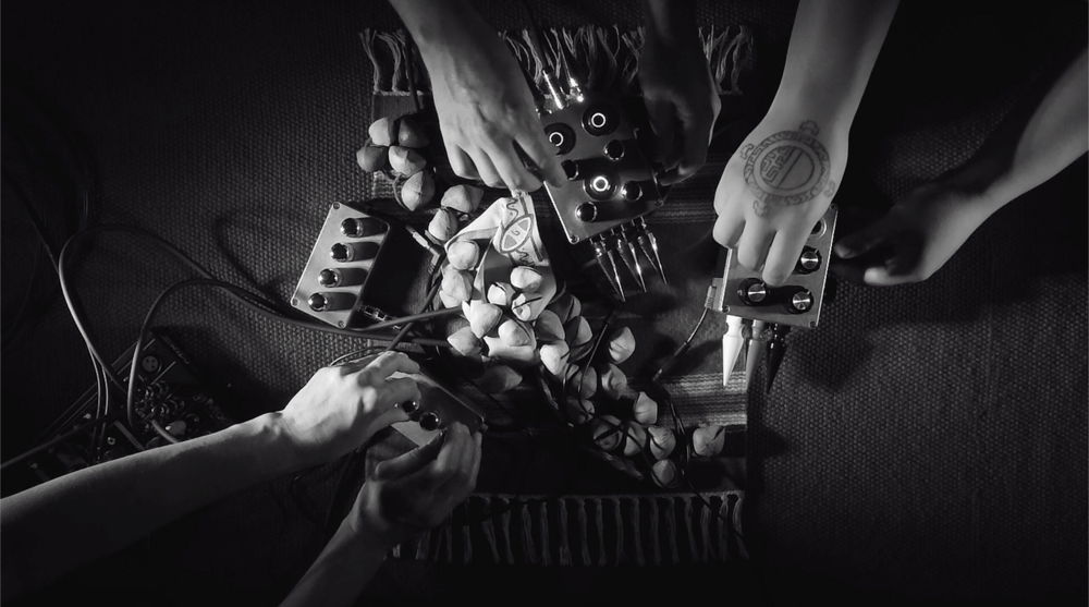 The hands of three people toying with wires of what appears to be electronic music  synthesizers shot from a birds-eye view angle in black and white.