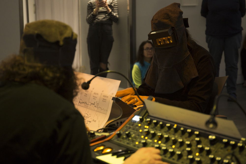 In a studio space, a performer playing the keyboard faces away from the camera and looks down at musical notation. Facing the camera is another musician whose face is covered by a mask with a screen attached to it, showing glinting electronic lights.