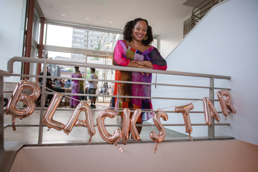 Maori Karmael Holmes poses smiling on a stairway balcony. She stands behind a railing hung with shiny inflatable bubble letters that spell out “Blackstar”.