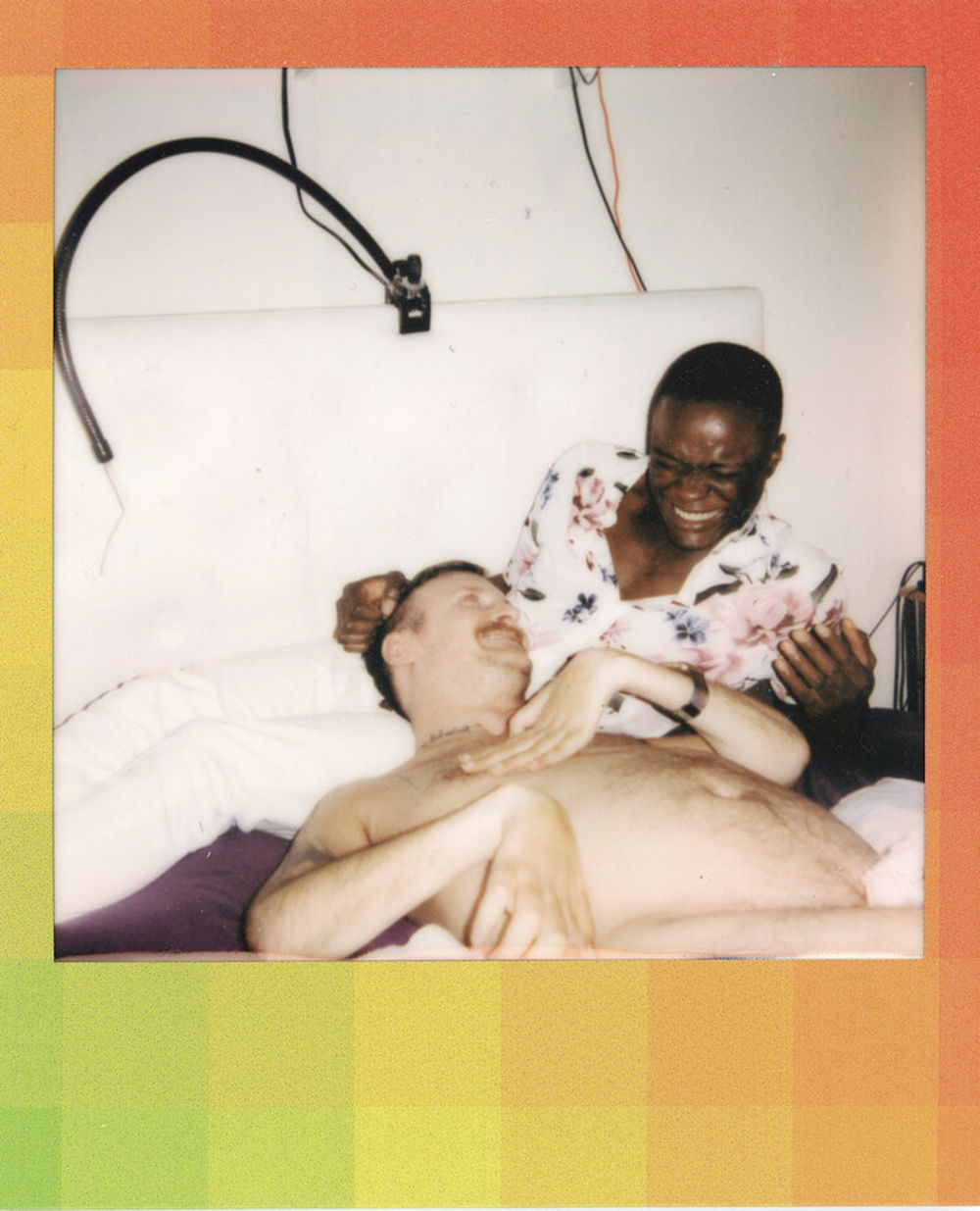 A brightly lit photograph of a shirtless White person laying in the lap of a Black person. Both figures' features are somewhat obscured by the brightness of the photograph, however, their relaxed poses and candid smiles are friendly and intimate. A multicolored border surrounds the photograph, a dreamy composition of green, orange, and yellow pixelated squares creating a soft gradient.