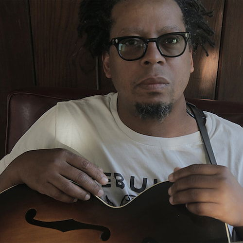 A portrait of a brown-skinned man sitting with his hands resting on a 1951 Gibson ES-150 electric guitar. He sits in shadow in front of a wood panelled wall, with specks of sunlight punctuating the image. He looks out at the viewer from behind black horn-rimmed glasses with an expressionless face. He has short dreadlocked hair and a small chin beard.