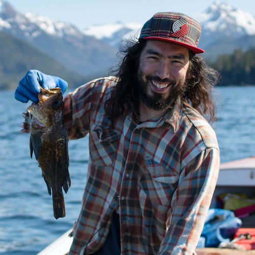 A young Yup'ik man smiles holding up a fish as he stands in a red skiff with blue water and snow-capped mountains in the background. He has dark long hair and a beard and is wearing a plaid shirt.