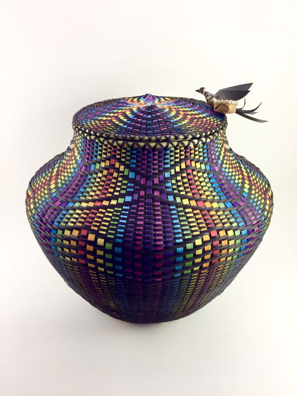 Photo of a brightly colored basket, with rainbow stripes woven through intricate patterns of black strips. A tiny woven bird rests on one edge of the basket lid.