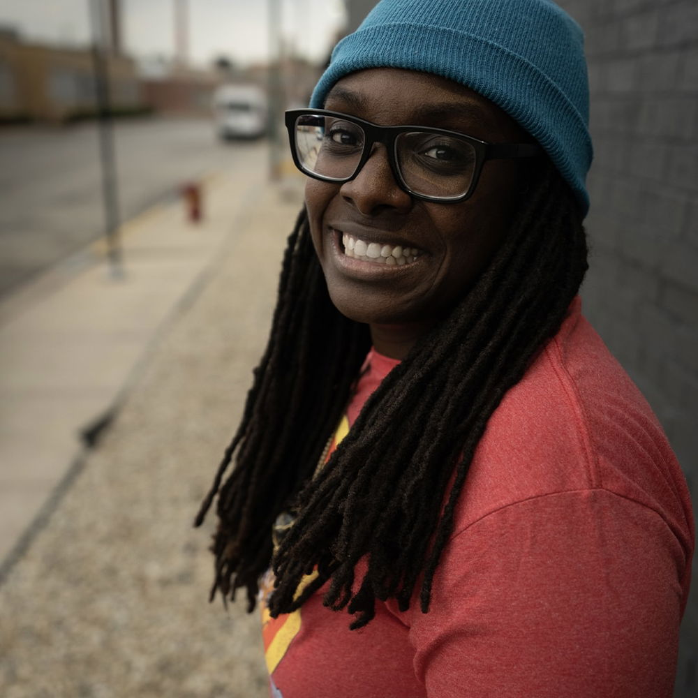 A black woman stands grinning against a black wall with the street out of focus behind her. She wears glasses, a red T-shirt, and a blue hat.