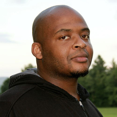 A headshot of a bald Black man wearing a black hoodie. <span style="font-weight: 400;">He looks towards the viewer, with a stoic expression</span>. Behind him are glimpses of trees and mountains.