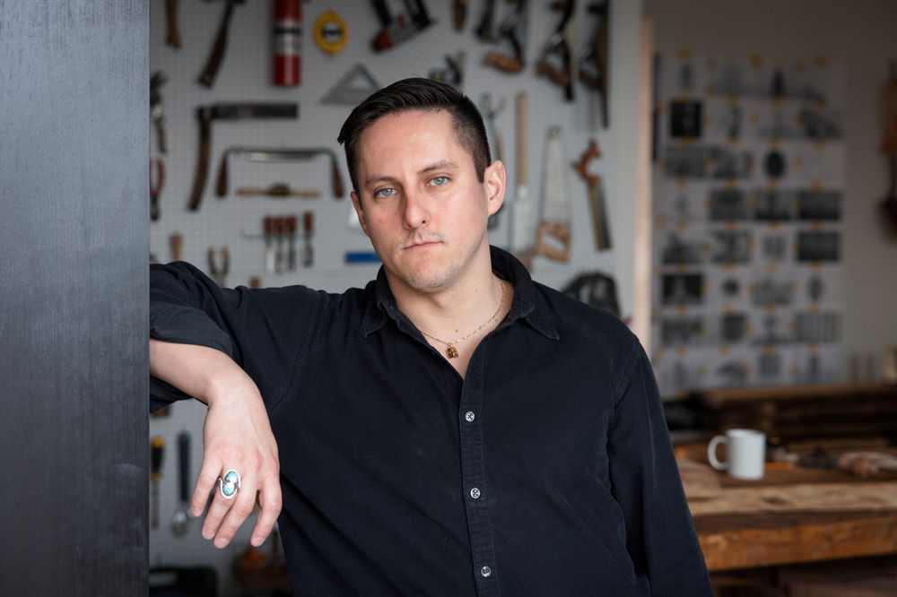 Raul, a man with light skin, green eyes, and short, black hair combed to the side, leans against a black bookshelf and gazes into the camera. Behind him is a pegboard hung with an array of tools.