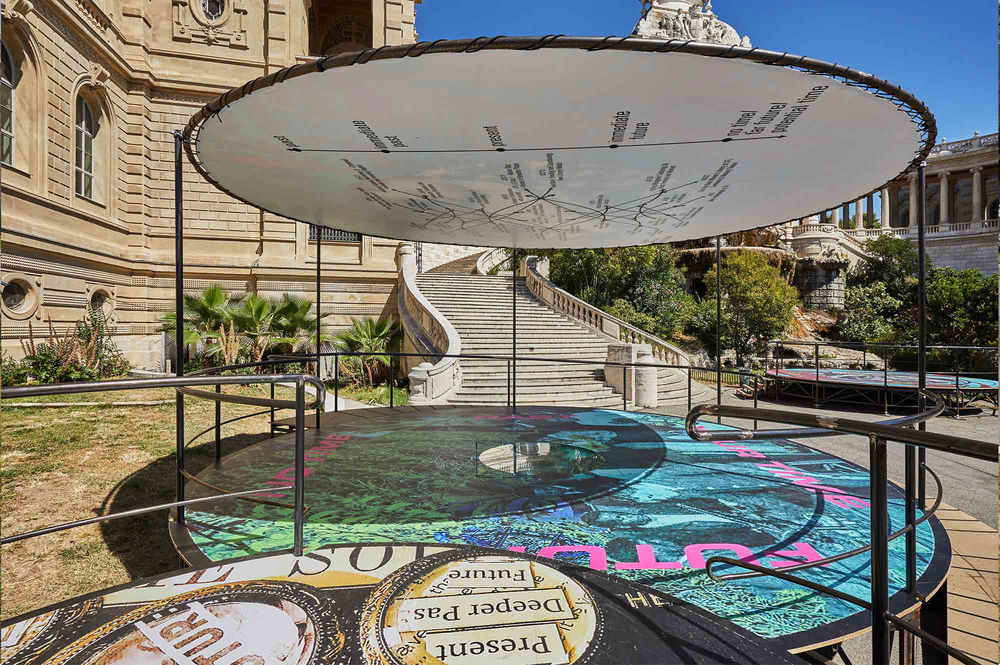 Outside the Palais Longchamp there is a cylindric stage interlocking with another cylindric stage. Both of the circular platforms have graphic text and digital textures patterning the floor.