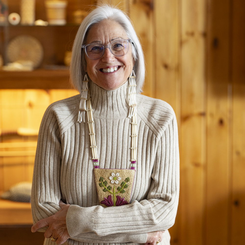 A woman with silver hair wearing eyeglasses, a cream-colored turtle-neck sweater and beaded jewelry poses with her arms crossed in a room with warm wooden paneling. She smiles widely.