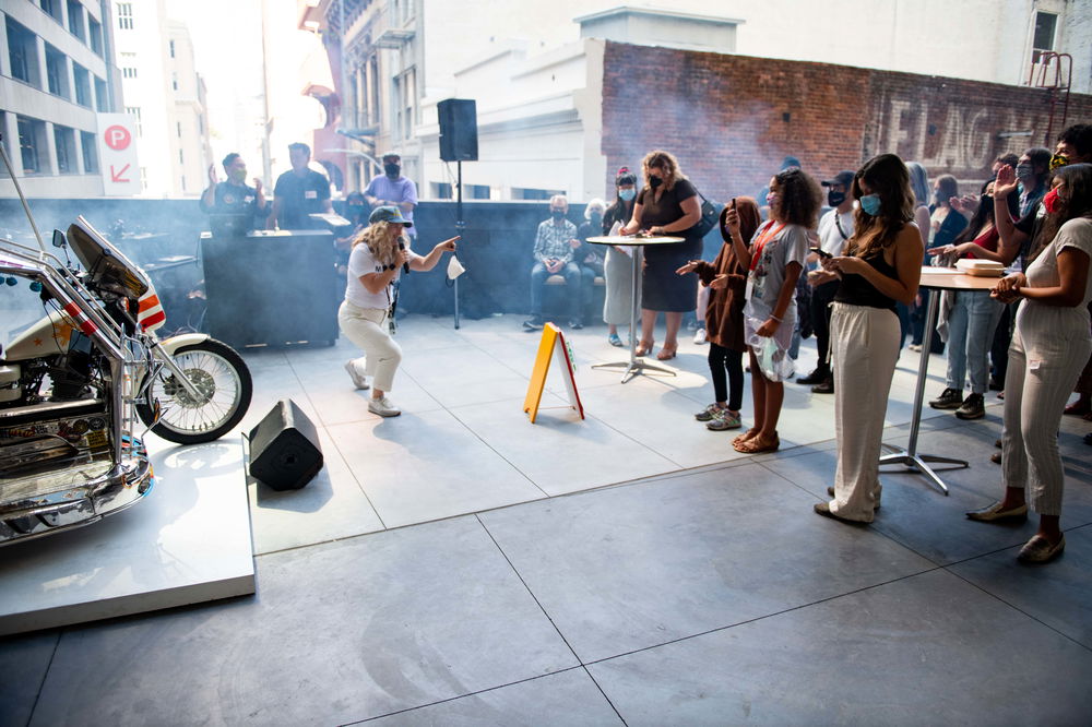 A woman wearing a blue cap, white t-shirt, white pants, and white sneakers, with blonde hair that falls past her shoulders, is holding a microphone and standing in a crouched position, pointing toward an audience of people who are standing in a crowd in front of a brick wall. She is singing in front of a Philippine traysikel (motorized-covered sidecar).