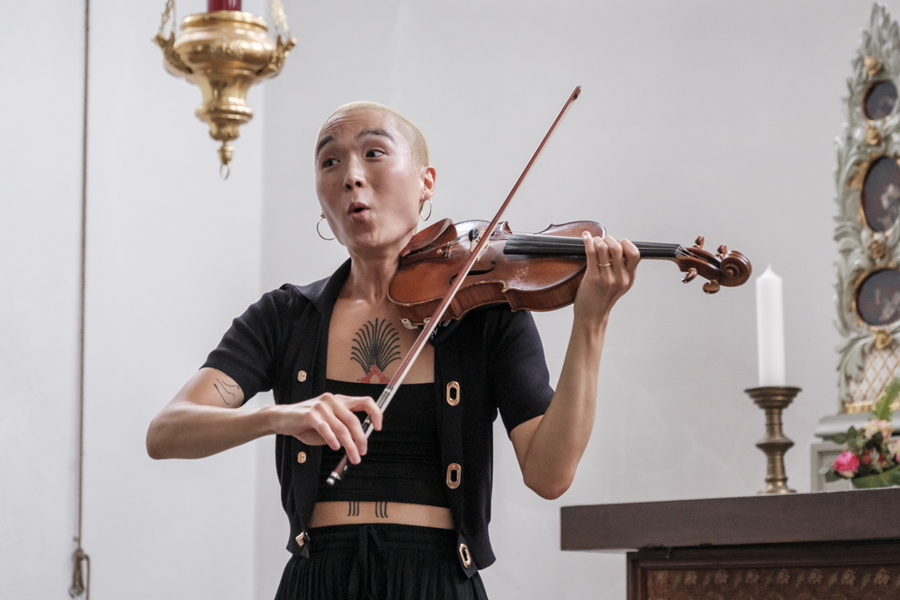 A person of Asian descent with a shaved head plays the violin. She is making an O-shape with her mouth, suggesting she may be singing or reacting to something with joy. She wears a black outfit with gold buttons that reveals portions of a large tattoo that covers the length of her torso. Behind her, to the right, is a mantlepiece with flowers and a large pillar candle. A gold lantern hangs above her to the left.