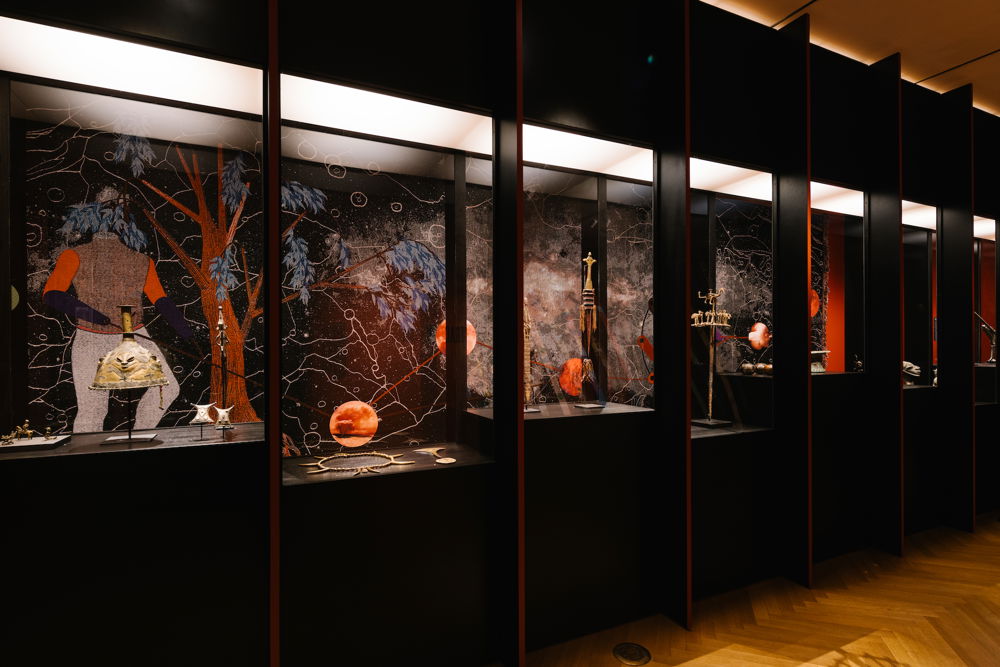 A wall of dark display cases divided into vertically-oriented rectangular sections. Each section displays warm metallic objects that resemble jewelry, figurines, or vessels. On the wall behind the objects is a mural depicting a figure wistfully standing behind a tree with red bark and blue leaves. The background of the mural is a moody black backdrop with constellations of white circles and webbing lines.