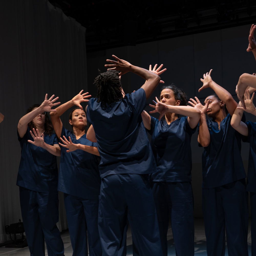 A group of dancers are dramatically lit on a stage. They wear matching dark blue scrubs and imitate the motions of the man standing in front of them. He faces the dancers, away from the camera, with his arms and hands shielding his face.