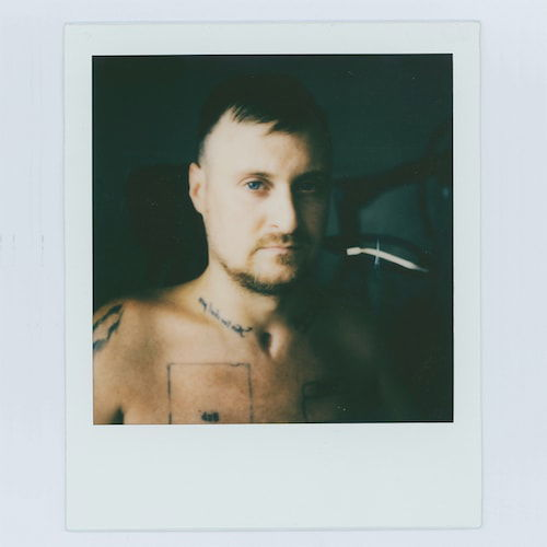 A Polaroid of a shirtless man seen from the shoulders up. His skin is lit in stark contrast, with the left half of his face in shadow. He has short brown hair and a goatee. His chest and shoulders are spotted with illegible tattoos.