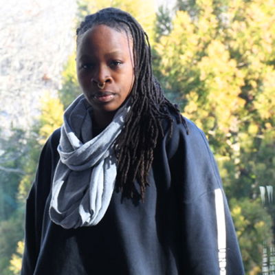Ni’Ja, a stylish Nonbinary Trans person with long locs draped over their left shoulder, stands in front of a window overlooking greenery and trees. In a dark sweatshirt and gray infinity scarf, they look directly at the camera.
