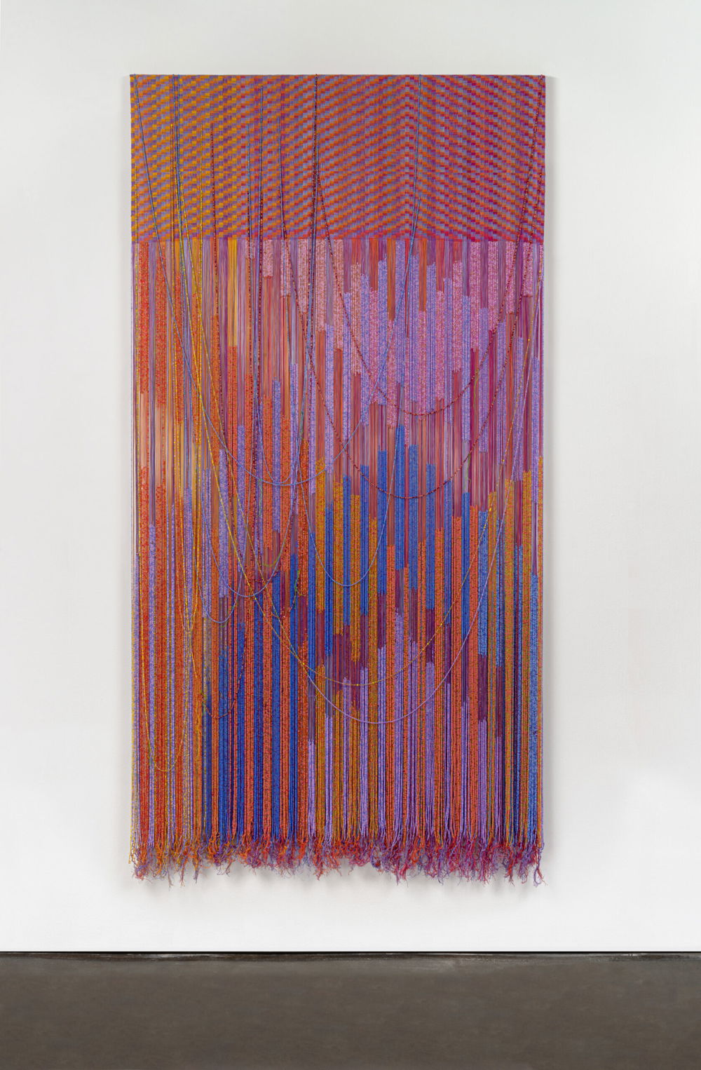 A vertical rectangular-shaped tapestry. The top quarter of the composition is a completed tapestry composed of geometric patterning in iridescent reds, oranges, and blues. The bottom three quarters of the composition are unwoven with intricately beaded strands of thread that create a vibrating pattern of intersecting chevrons in saturated reds, oranges, pinks, purples, and blues. A few loose strands are wisped upward creating thin U-shaped lines.