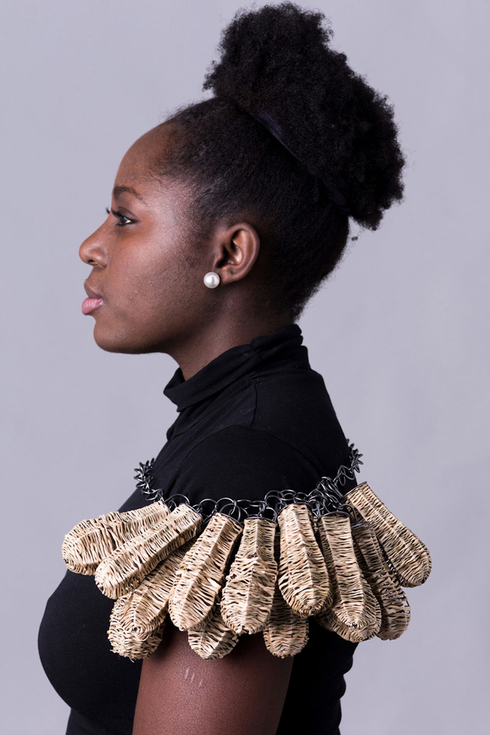 A dark-skinned person is photographed against a gray backdrop. They are captured from their left side profile and are dressed in a plain black T-shirt. A sterling silver corded necklace resembling a telephone cord rests just beneath their shoulders. On the left side of the wire, a cluster of oblong woven sea grass and 14 nu-gold pendants hang and rest over their shoulder.
