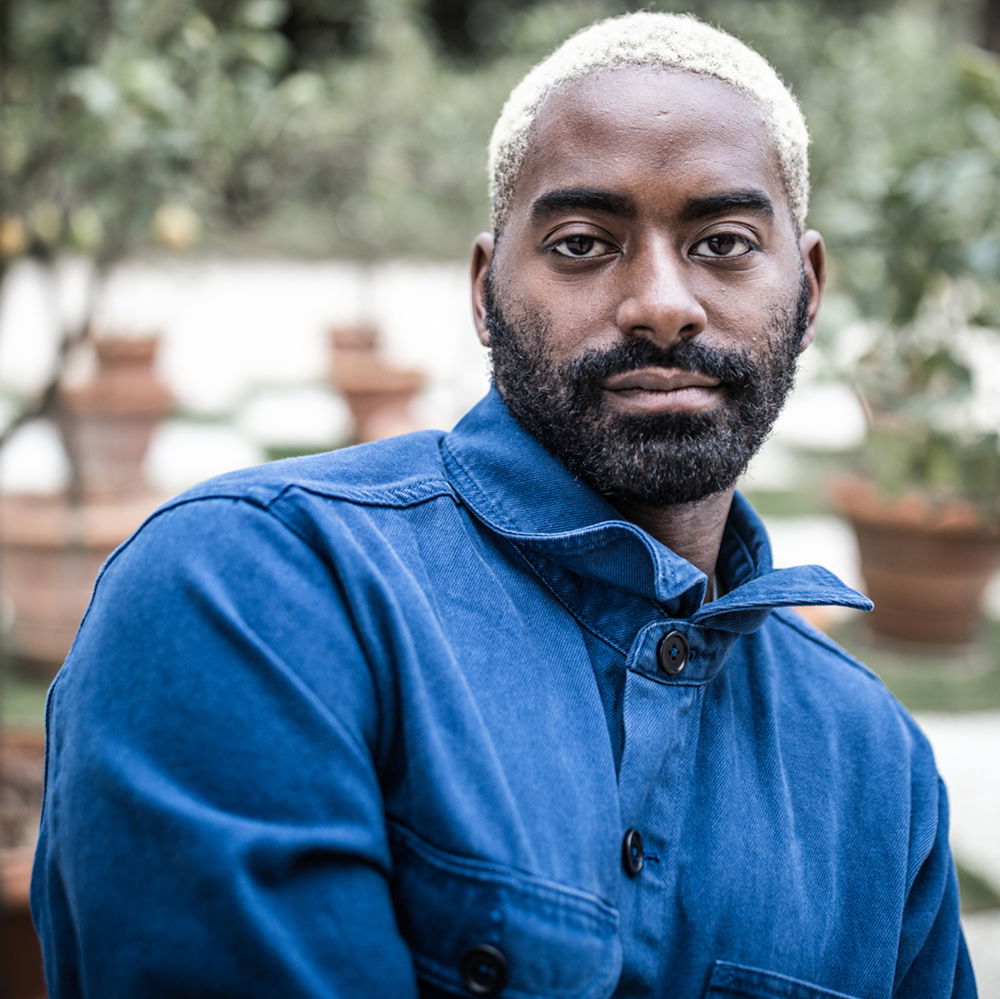 A dark-skinned Black man with a beard and blonde hair stands wearing a bright-blue worker jacket in front of a garden.