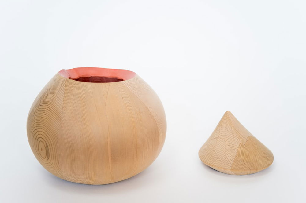 Photograph of a rounded wooden vessel with the grain visible on its surface. The lid, made of the same type of wood, comes to a conical point and rests alongside it. The inside of the vessel is painted a salmon color.