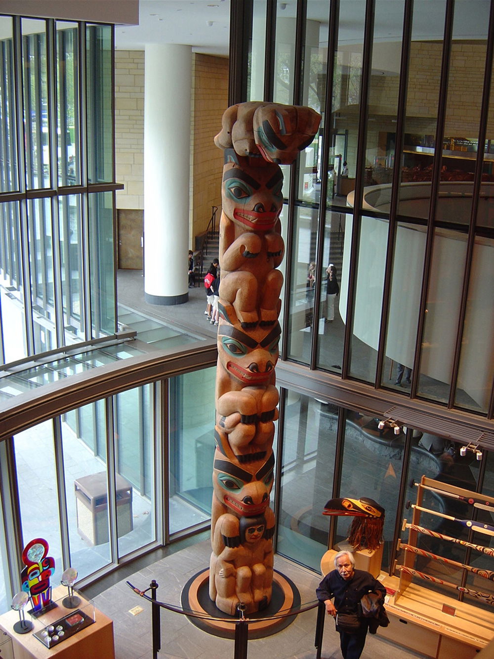 Nathan P Jackson stands below a towering totem pole on display in the Smithsonian National Museum of the American Indian. The totem features Kaats who is at the bottom being held by the mama bear, his wife. Going up the pole, the three bear cubs are shown.