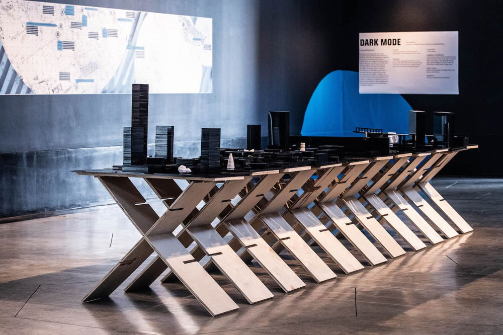 Photo of an installation in a space with black-painted walls. A long table supported by x-shaped frames is covered with a model-sized city of black buildings. A sign reading “Dark Mode” hangs behind the table.