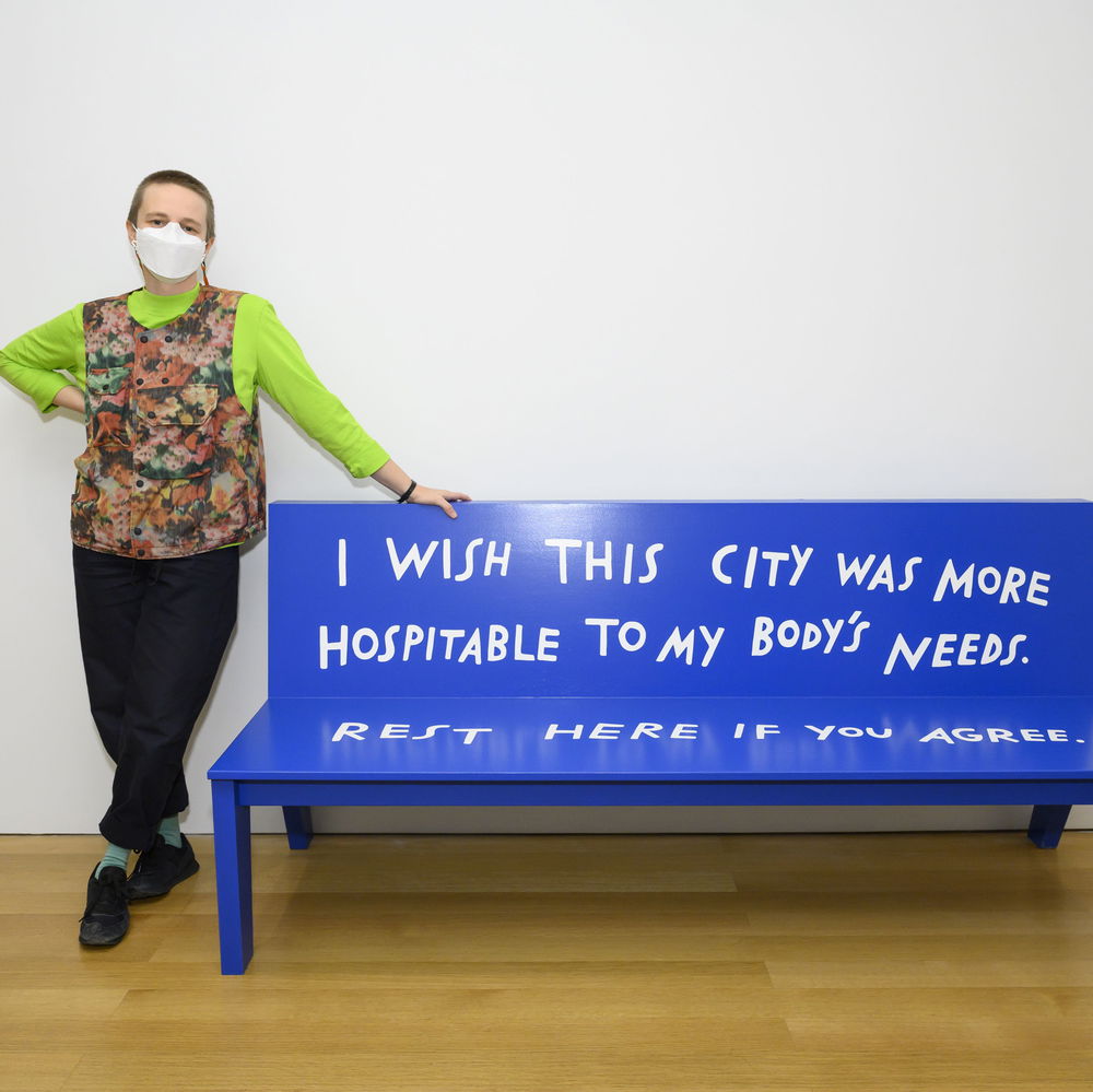 Finnegan Shannon, a white person with a buzz cut, posed next to a bright blue bench. Their handwriting on the bench reads, "I wish this city was more hospitable to my body's needs. Rest here if you agree." They wear a KF94 mask, a fluorescent lime shirt, and a floral fisherman-style vest.