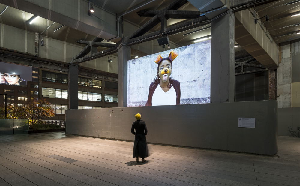 Projected onto a concrete industrial wall, Lex Brown, a Black woman in a costume dog nose and dog ears, sticks her tongue out. A person stands below the projection and watches.