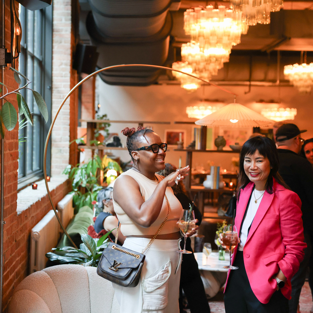 Two guests hold glasses of wine and converse at a lounge with an arched lamp and bookshelves behind them. Light from a wall of windows on their left softly illuminates them.