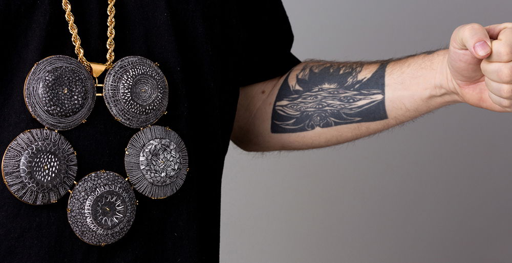 A cropped photograph of a person’s chest and arm wearing a black T-shirt with a gold-plated brass, copper, and enamel necklace. The necklace boasts five intricately detailed medallions. On the forearm a black rectangular tattoo can be seen with details of multiple eyes.
