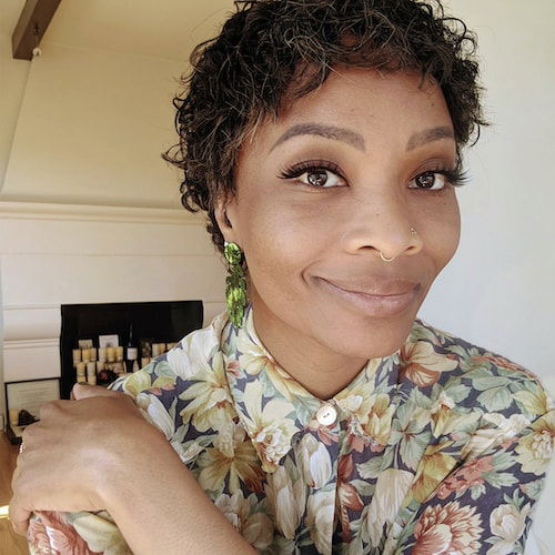 A headshot of a nonbinary artist with brown skin, large dark eyes and short curly hair smiles at the camera. Their right arm is crossed over their left shoulder and rests on their floral shirt. A grouping of candles, representing their ancestors, can be seen behind them.