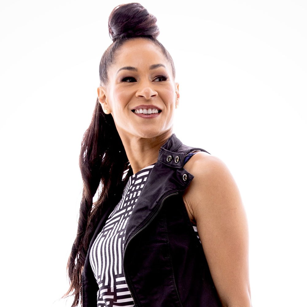 A woman of color with long hair and top knot bun wearing a black vest and black-and-white patterned top smiles and looks to her left against a white background.