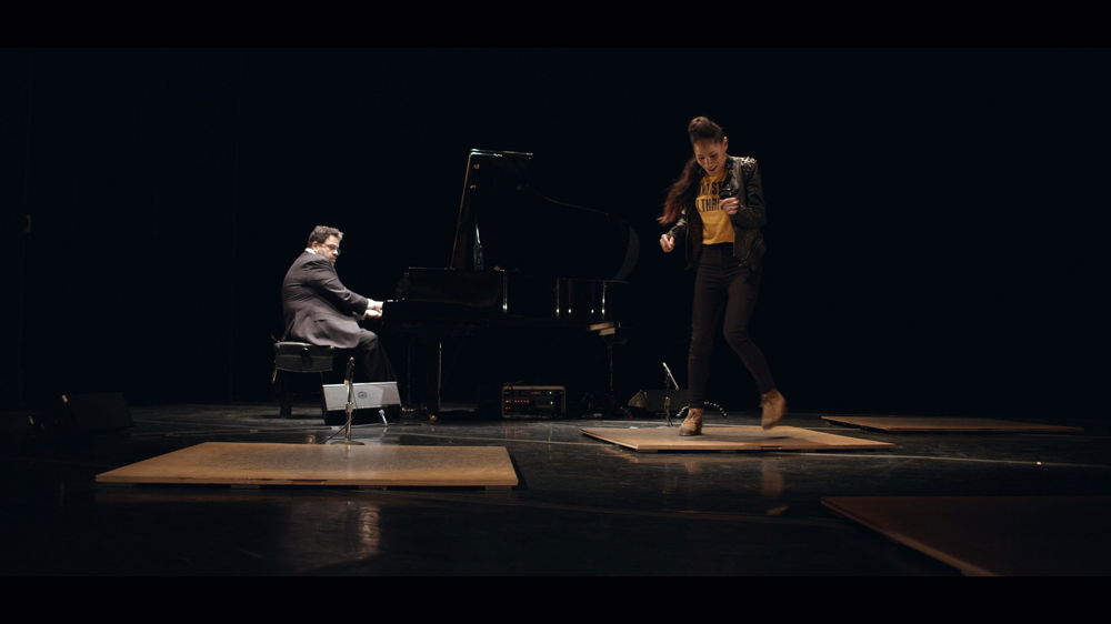 A performer with long, dark brown hair dances joyfully on a dark stage in front of a musician playing a baby grand piano.