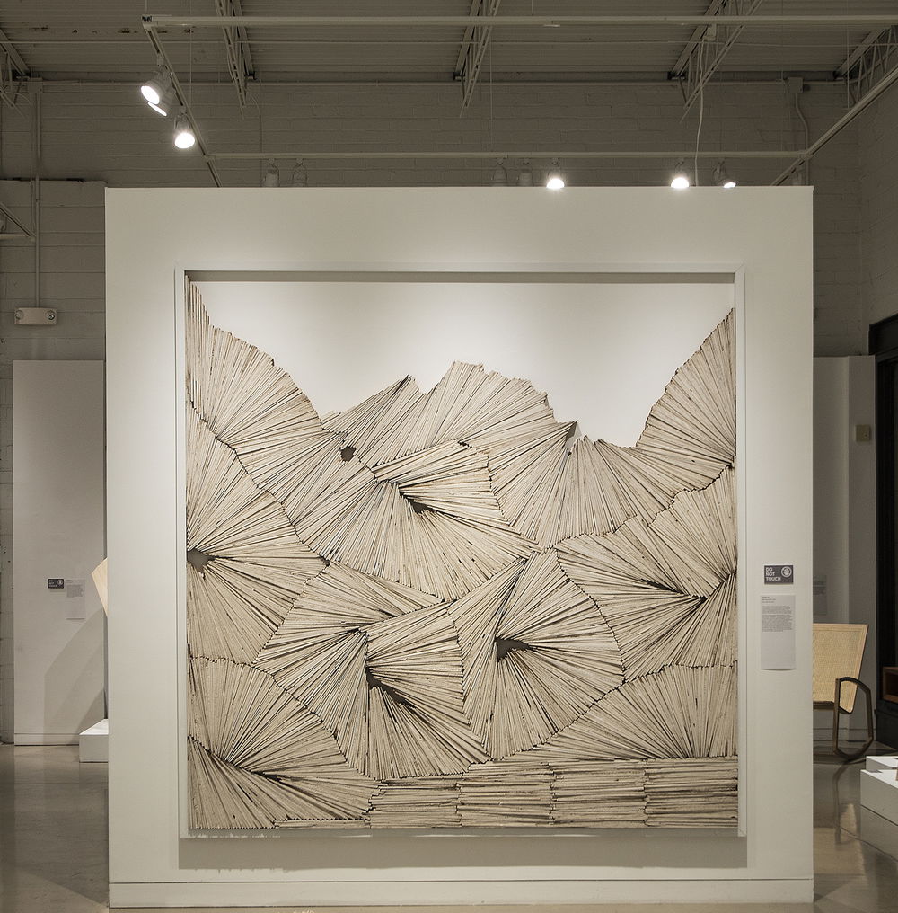 Thin, flat pieces of off-white painted wood are stacked together on a white background in shapes that look like stacks of books at the floor of the piece, then groups of cells in the middle. The rows of wood at the top of the stack are evocative of sharp craggy mountains.