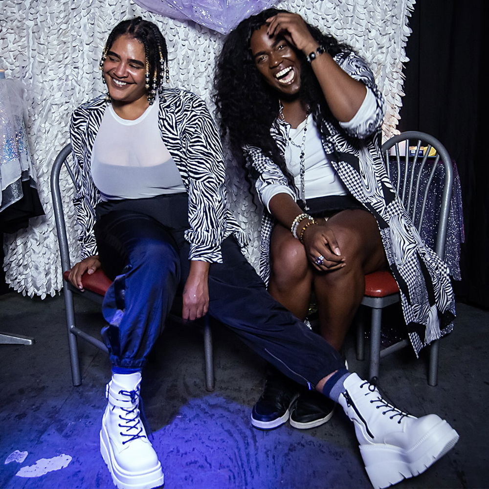 A photograph of two Black people, indee mitchell and Nathalie Nia Faulk, laughing and looking fantastic in front of a sparkling purple-and-white backdrop. They are both wearing black and white. mitchell stretches their leg and white platform boot out in front of them. Faulk is bent to the side with a wide grin, arm raised to touch their forehead.