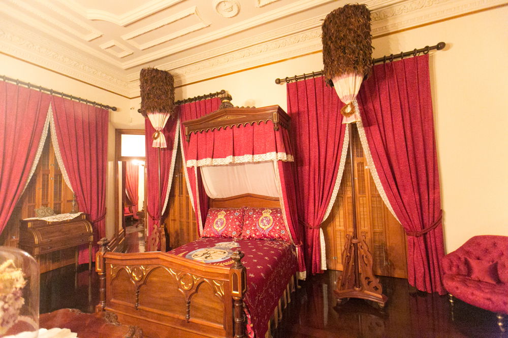 A regal bedroom with polished wooden furniture and lush burgundy curtains and bedding. Two identical thin, towering sculptures with large conical tops covered in brown feathers flank the bed.