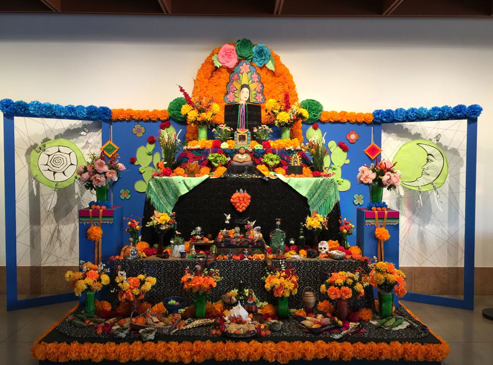A multi-level altar covered with many artifacts, flowers, and textiles. An image of the goddess Ix Chel is located at the highest point, surrounded by hand-made paper flowers. The altar is framed on the left and right by images of the Sun and Moon shields.