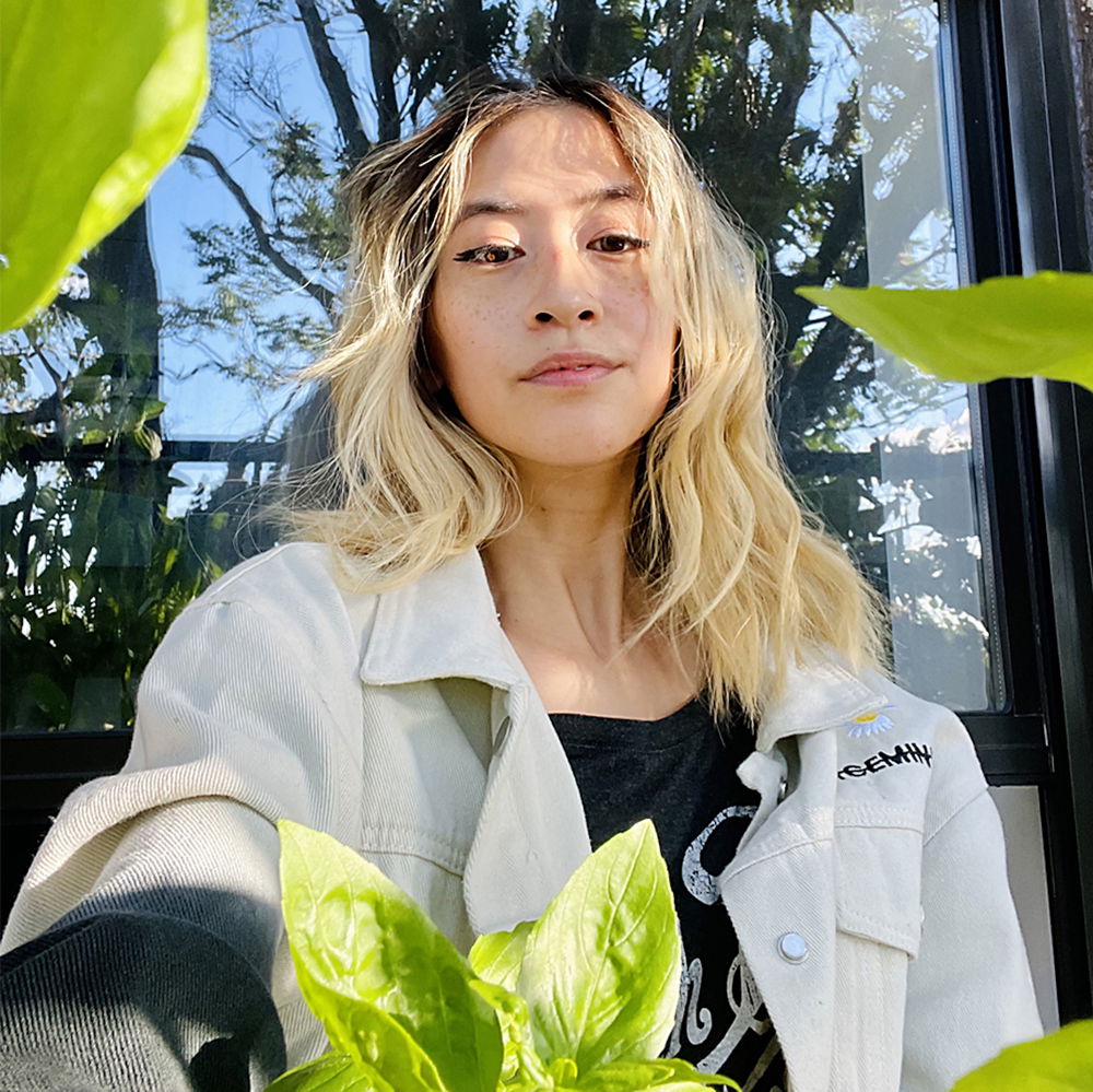 A thin Taiwanese femme with bleached blonde hair and black winged eyeliner sits on their fire escape surrounded by basil leaves in their garden.