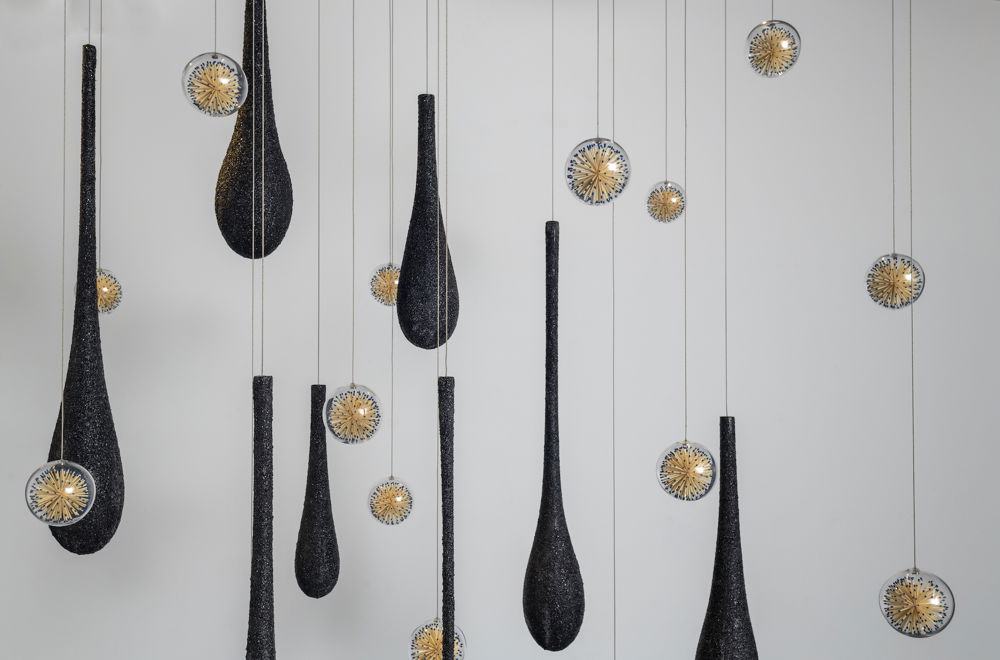 Glass sculptures suspended in air resemble large black droplets of water and clear orbs filled with matches. The matches are unused and are arranged into a flowering sphere shape.