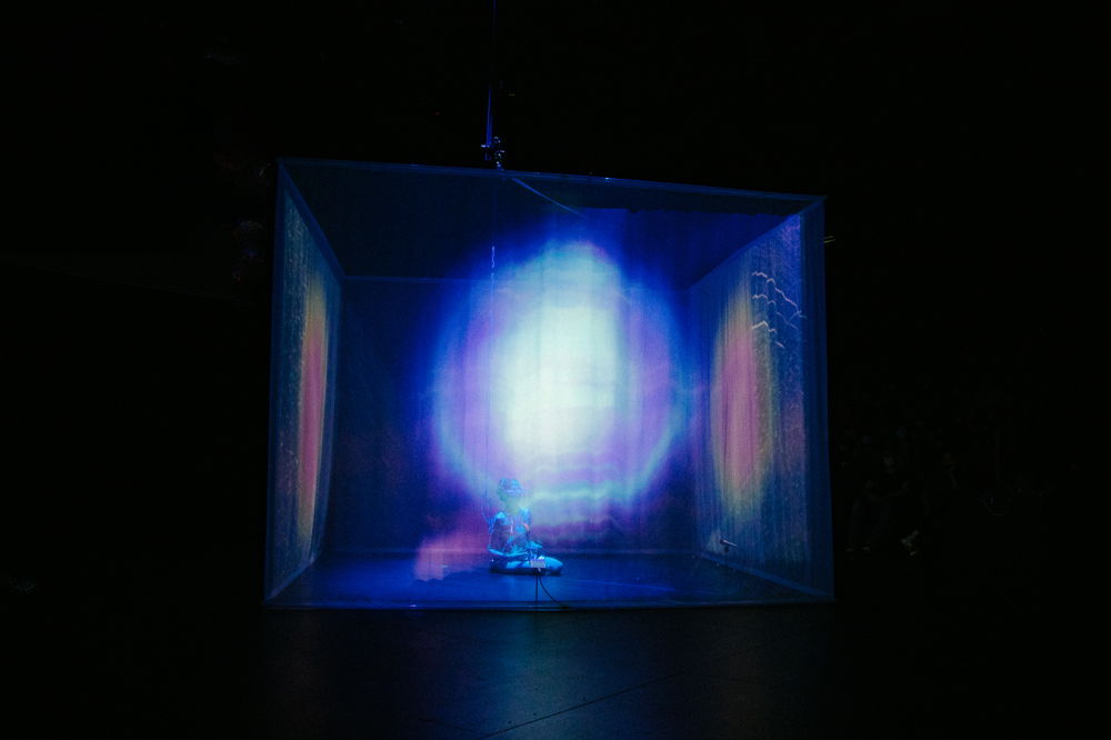 A thin ghostly figure sits in a cube made of translucent scrims. The room is dark except for the spectral orbs projected onto the screen, which glow an icy blue, dim orange, and unnatural green-yellow.