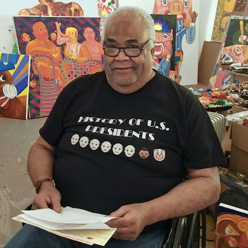 A portrait of a man in a studio, surrounded by paintings. The man has brown skin and short grey hair. He sits in a black wheelchair, looking up at the viewer from behind black-rimmed glasses. He wears an amused expression and holds a pile of papers. He wears a black t-shirt that reads "History of US Presidents" with several "White man" emojis, a "Black man" emoji, and a "clown" emoji.