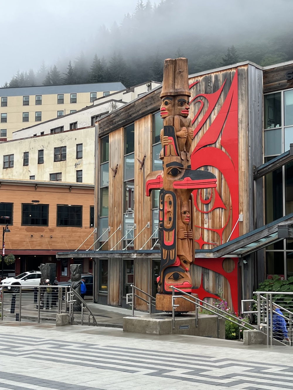 A tall totem pole in an urban setting on a foggy day. The long wooden pole is ornately carved with a cast of stylized characters. Each character overlaps one another and is painted in a color palette of red and black.