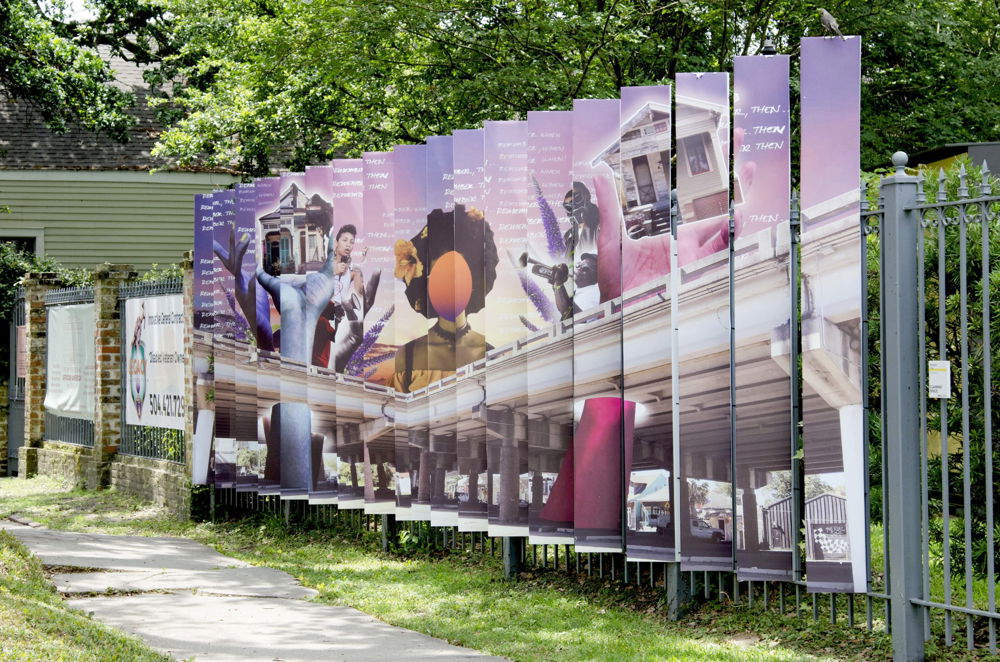 Tall vertical panels are affixed to an iron gate creating the illusion of one long rectangular banner. On the banner are collaged photographic and graphic images of an overpass, hands reaching up to the sky holding houses, and a portrait of a faceless woman with medium-brown skin and a hibiscus flower in her hair.