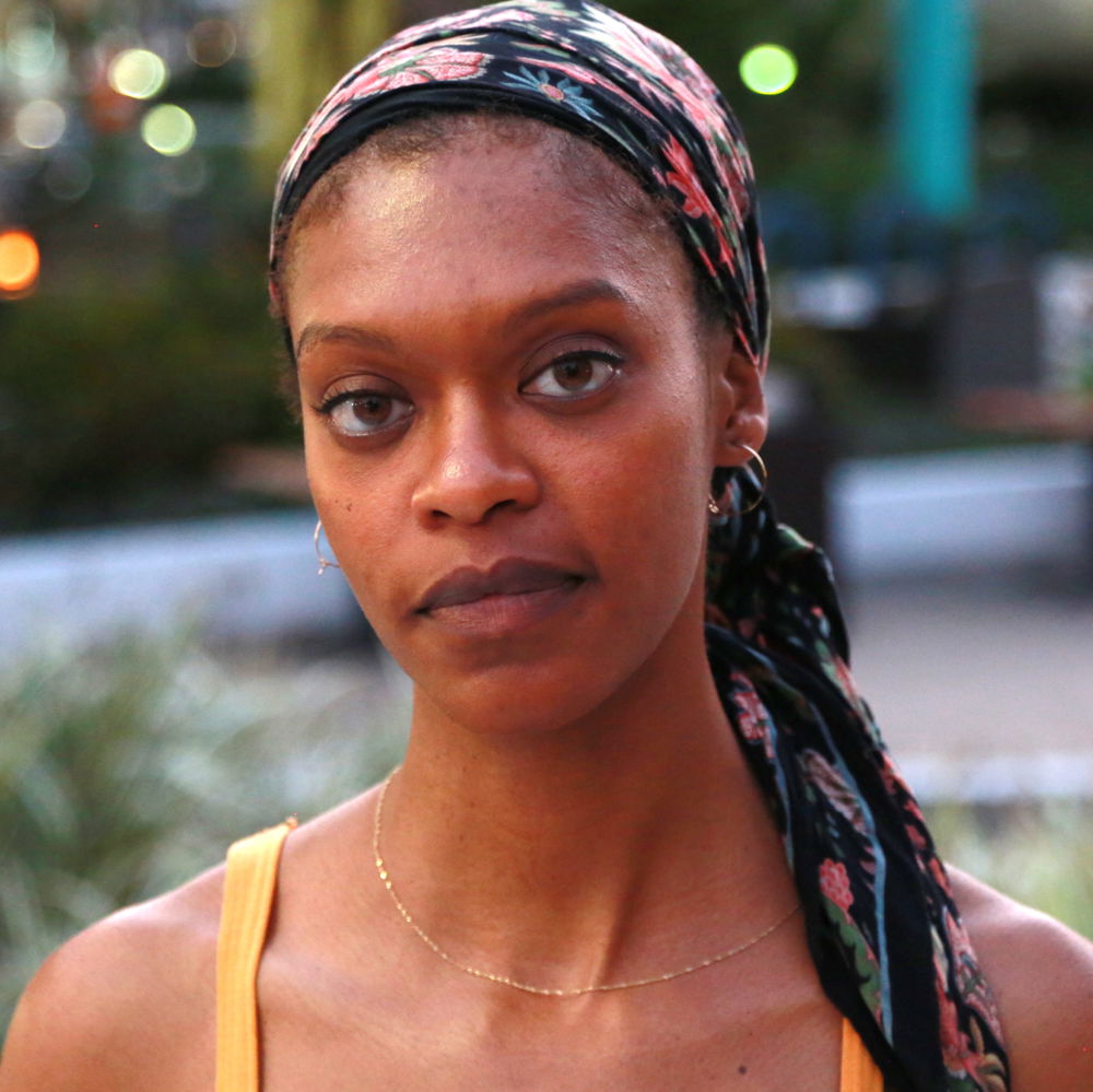 Faren, a Black person adorned with a floral headscarf, looks intently at the camera while standing amongst a public square at dusk; polychromatic lens flares abound.